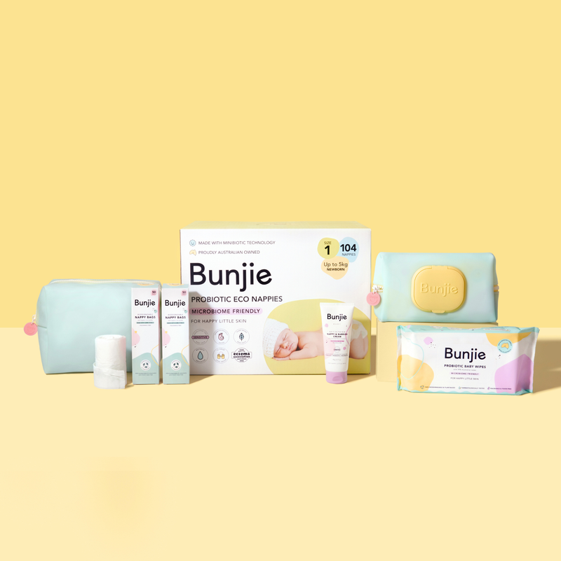 A collection of baby care products from the brand Bunjie arranged on a solid pale yellow background. The products include a light blue zippered pouch, a white bottle, two small boxes, a tube, a larger white box, a pastel blue pouch with a yellow lid, and a colorful box of wipes.